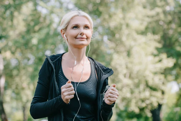 Beautiful mature sportswoman looking away while running in park and listening music in earphones — Stock Photo