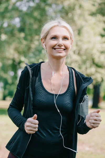 Cheerful mature sportswoman jogging in park and listening music in earphones — Stock Photo
