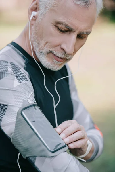 Красивый зрелый спортсмен, использующий смартфон в футляре для повязки и слушающий музыку в наушниках — стоковое фото