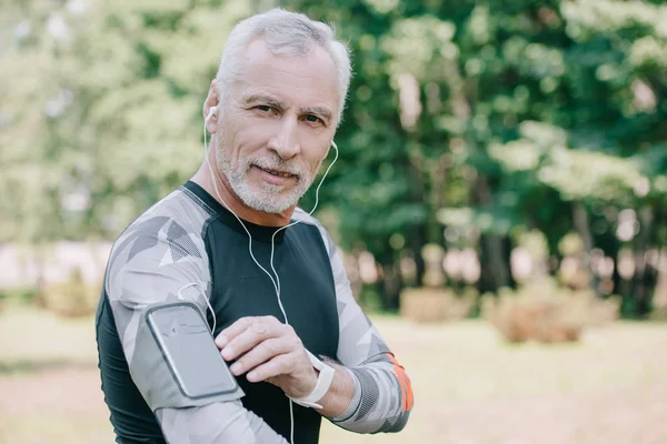 Beau sportif mature regardant la caméra tout en utilisant un smartphone dans le boîtier du brassard et en écoutant de la musique dans les écouteurs — Photo de stock