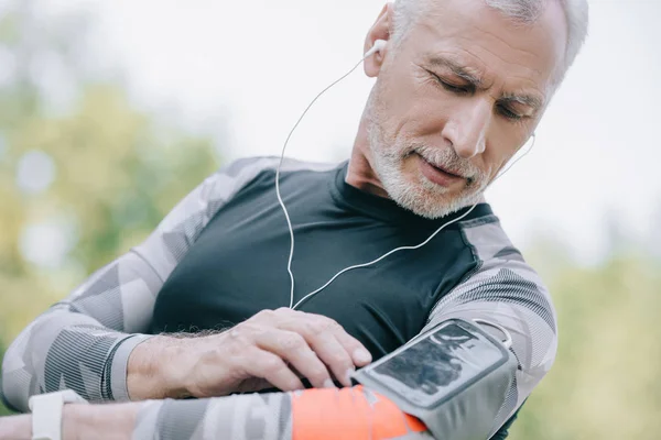 Guapo deportista maduro usando smartphone en estuche de brazalete y escuchando música en auriculares - foto de stock