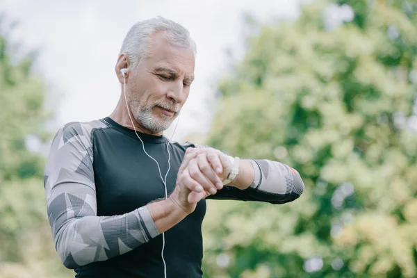 Apuesto deportista maduro escuchando música en los auriculares mientras mira el rastreador de fitness - foto de stock