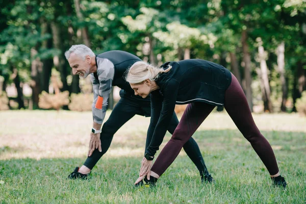 Positive, mature sportsman and sportswoman stretching in park together — Stock Photo