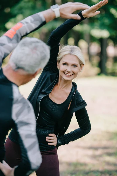 Усміхнена спортсменка прогрівається в парку біля зрілого спортсмена — стокове фото