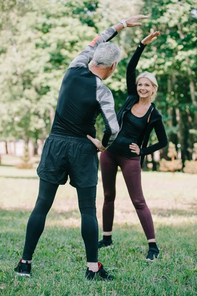 Back view of mature sportsman warming up near attractive, smiling sportswoman in park — Stock Photo