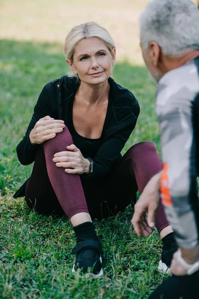 Rückansicht eines Sportlers, der neben einer verletzten Sportlerin steht, die auf dem Rasen sitzt und Bein berührt — Stockfoto