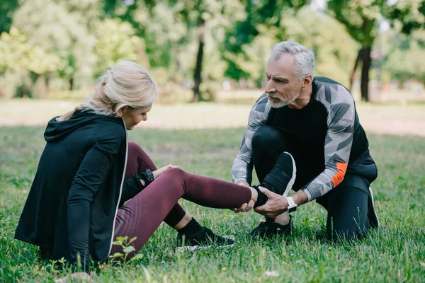 Älterer Sportler berührt verletztes Bein einer Sportlerin, die auf Rasen im Park sitzt — Stockfoto