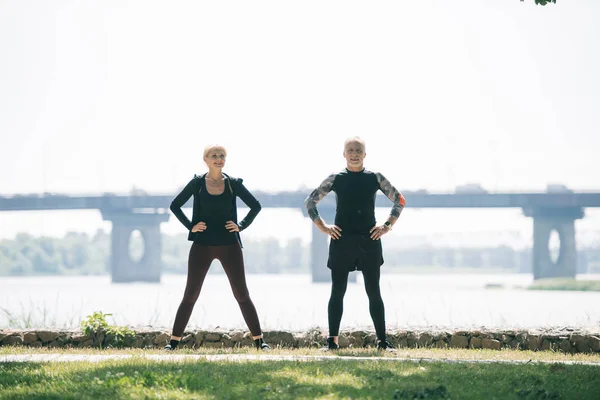 Mature sportsman and sportswoman looking at camera while training on riverside in park — Stock Photo