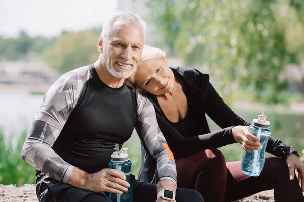 Positiver, reifer Sportler und Sportlerin sitzt auf Brüstung im Park und hält Sportflaschen in der Hand — Stockfoto