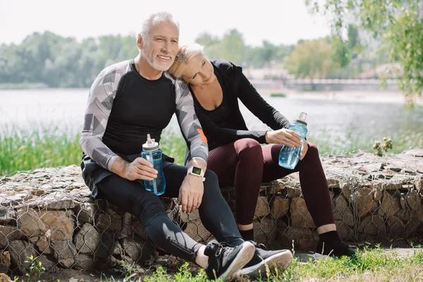 Улыбающийся взрослый спортсмен и спортсменка, сидящие на парапете в парке и держащие в руках спортивные бутылки — стоковое фото