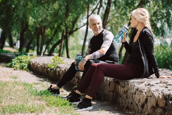 Sportif mature souriant et sportive assise sur la chaussée dans le parc et tenant des bouteilles de sport — Photo de stock