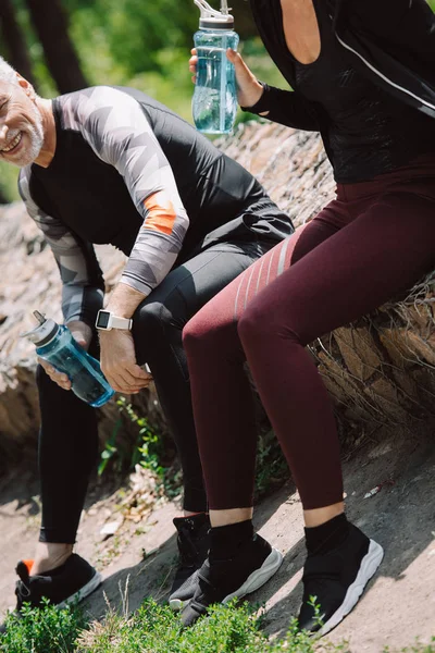 Ausgeschnittene Ansicht eines Sportlers und einer Sportlerin, die auf einem Bürgersteig im Park sitzen und Sportflaschen in der Hand halten — Stockfoto