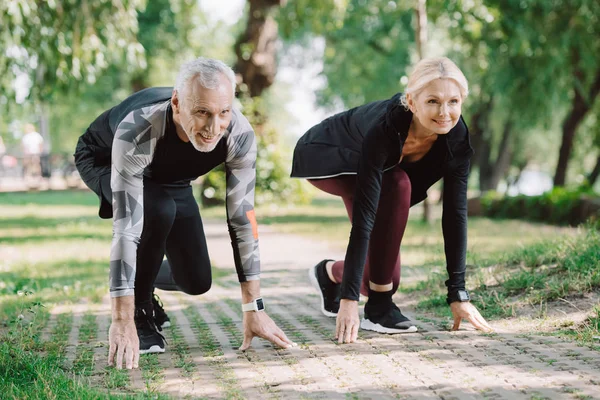 Cheerful mature runners standing in start positing on pavement in park — Stock Photo