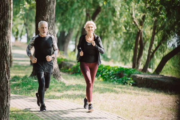 Mature, smiling joggers running together on pavement in sunny park — Stock Photo