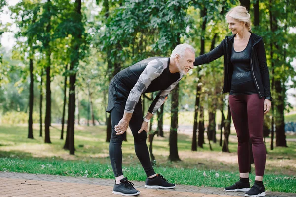 Tired mature sportsman standing near smiling sportswoman in park — Stock Photo