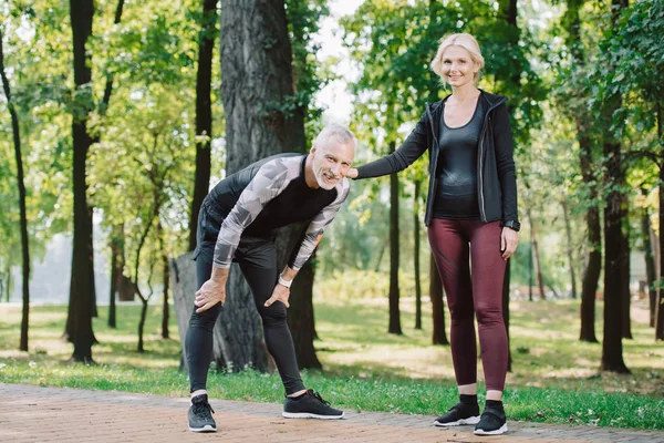 Уставший взрослый спортсмен и улыбающаяся спортсменка, смотрящая вместе, стоя в парке — стоковое фото