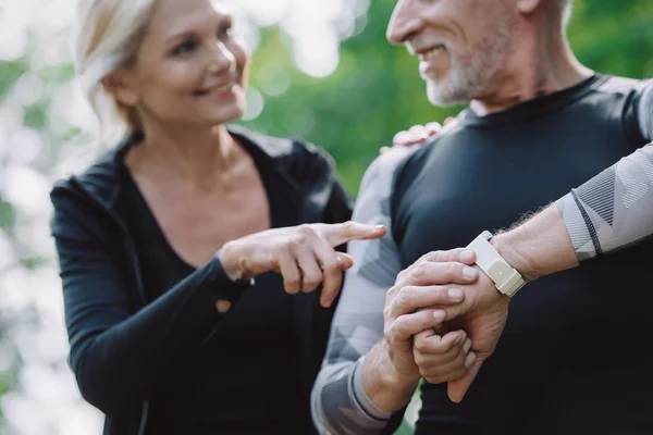 Selective focus of smiling sportswoman pointing at smart watch on hand of mature sportsman — Stock Photo