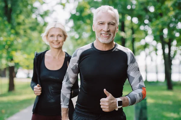 Cheerful mature sportsman showing thumb up while jogging in park together with smiling sportswoman — Stock Photo