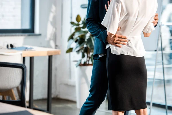 Cropped view of businessman hugging businesswoman while standing and flirting in office — Stock Photo