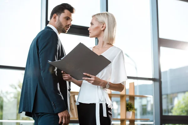 Guapo hombre de negocios mirando atractiva mujer rubia sosteniendo carpeta en la oficina - foto de stock