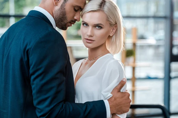 Guapo barbudo hombre con los ojos cerrados abrazando atractiva mujer rubia mirando a la cámara - foto de stock