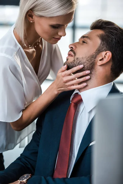 Enfoque selectivo de la mujer rubia apasionada tocando la cara del hombre guapo en la oficina - foto de stock