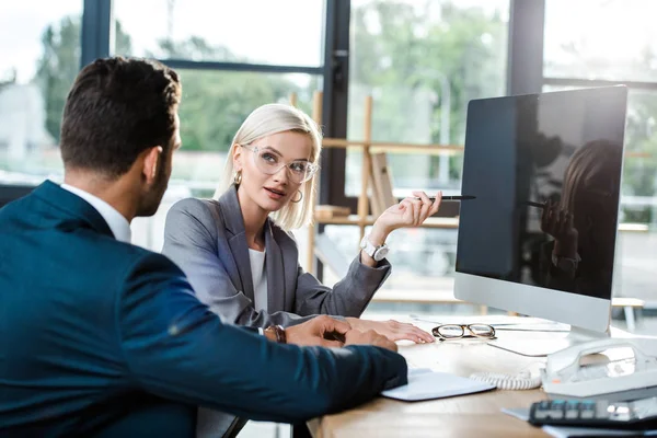 Selektiver Fokus attraktiver Geschäftsfrau in Brille, die Mitarbeiter in der Nähe des Computermonitors mit leerem Bildschirm anschaut — Stockfoto