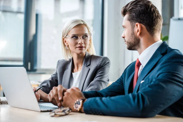 Enfoque selectivo de mujer rubia atractiva en gafas mirando hombre de negocios cerca de la computadora portátil - foto de stock