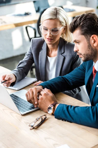 Attraktive Frau mit Brille hält Stift in der Nähe von Laptop neben bärtigen Mann im Büro — Stockfoto