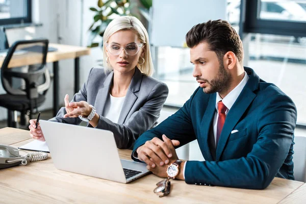 Attrayant femme dans des lunettes pointant avec le doigt à l'ordinateur portable près homme barbu au bureau — Photo de stock