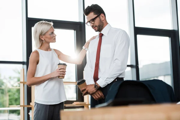 Selective focus of attractive blonde businesswoman looking at upset man in glasses holding paper cup — Stock Photo