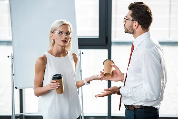 Atractiva mujer rubia y hombre guapo en gafas haciendo gestos mientras sostiene vasos de papel - foto de stock