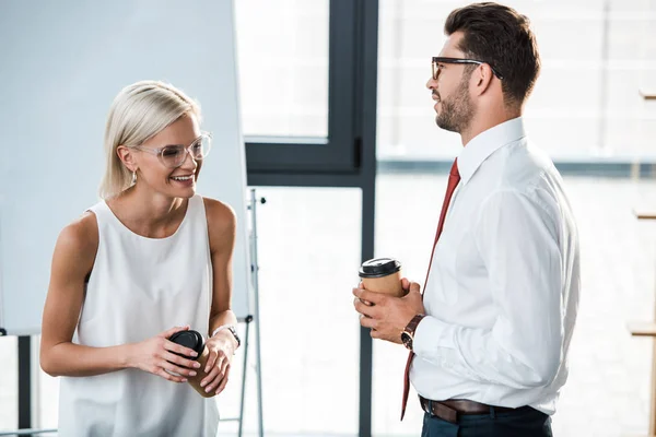 Guapo barbudo hombre de negocios sosteniendo taza desechable y mirando alegre mujer rubia en la oficina - foto de stock