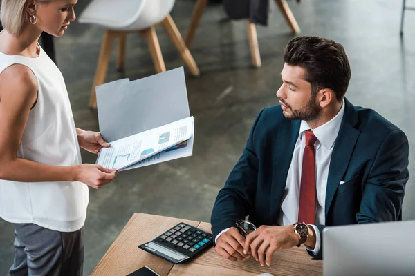 Vista aérea de la mujer de negocios rubia sosteniendo carpeta con gráficos y gráficos cerca de hombre de negocios barbudo en la oficina - foto de stock