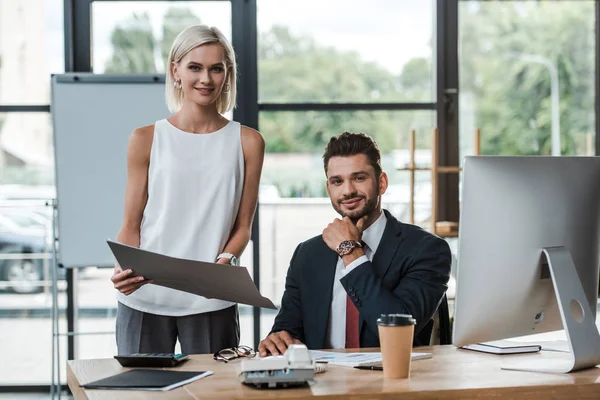 Selective focus of happy blonde businesswoman holding folder near cheerful bearded businessman in office — Stock Photo