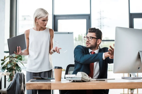 Selektiver Fokus einer unzufriedenen blonden Geschäftsfrau, die gestikuliert, während sie Ordner in der Nähe eines hübschen Mannes hält — Stockfoto