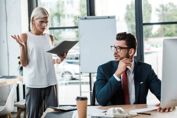 Selective focus of emotional blonde businesswoman gesturing while holding folder near handsome man — Stock Photo