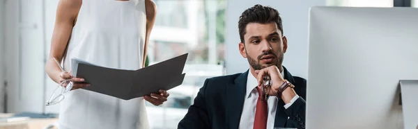 Panoramic shot of woman standing with folder near bearded man looking at computer monitor — Stock Photo