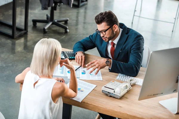 Selektiver Fokus eines gutaussehenden Geschäftsmannes mit Brille, der gestikuliert, während er Diagramme und Grafiken in der Nähe eines blonden Kollegen betrachtet — Stockfoto