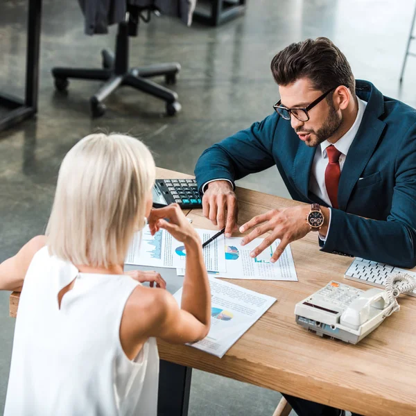 Enfoque selectivo de hombre de negocios en gafas gesto mientras mira gráficos y gráficos cerca de colega rubia - foto de stock