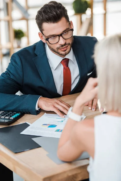 Handsome bearded man in glasses sitting near charts and graphs and looking at blonde woman — Stock Photo