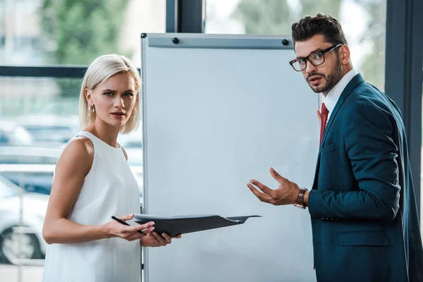 Attractive blonde woman holding folder near businessman in glasses gesturing in office — Stock Photo