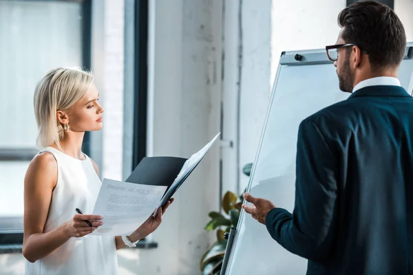 Attraente donna bionda che tiene la cartella e guarda i documenti vicino all'uomo barbuto gesticolando in ufficio — Foto stock