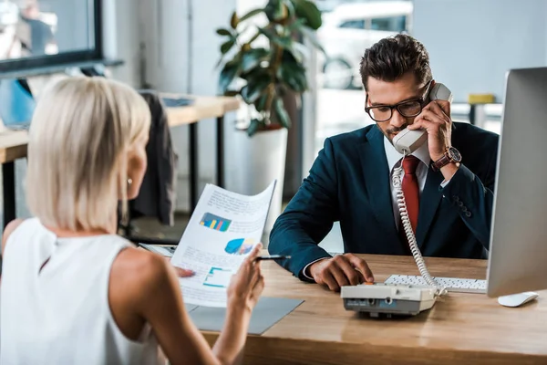 Selective focus of businessman in glasses talking on retro phone near blonde colleague looking at charts and graphs — Stock Photo