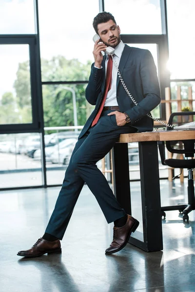 Hombre de negocios guapo con la mano en el bolsillo hablando en el teléfono retro en la oficina moderna - foto de stock