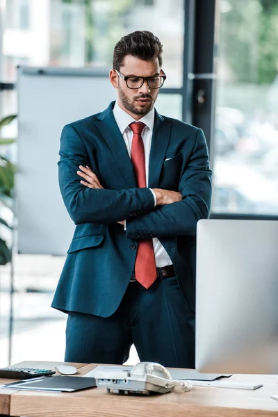 Seriöser Geschäftsmann mit Brille, der mit verschränkten Armen auf den Computermonitor blickt — Stockfoto