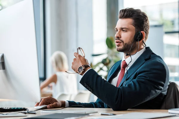 Selektiver Fokus des gut aussehenden Bedieners im Headset, der auf den Computermonitor blickt und eine Brille hält — Stockfoto