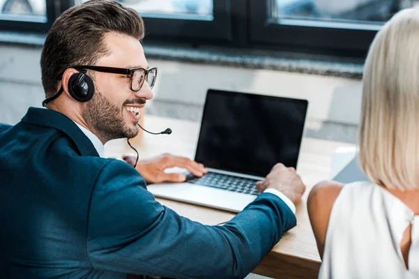 Enfoque selectivo de hombre alegre en gafas y auriculares mirando rubia compañero de trabajo en la oficina - foto de stock