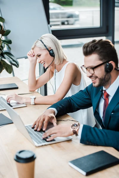 Enfoque selectivo de la mujer feliz en auriculares cerca de compañero de trabajo alegre usando el ordenador portátil mientras trabaja en la oficina - foto de stock