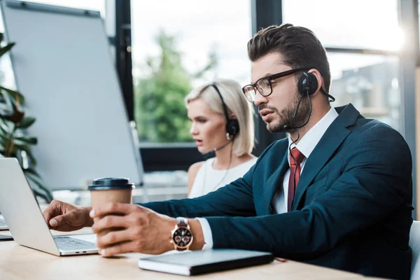 Selective focus of handsome operator in headset holding coffee to go near laptop and blonde colleague in headset — Stock Photo